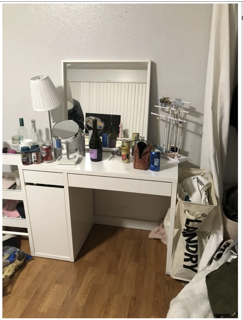 White vanity desk with mirror, lamp, jewelry stand, bottles and cans beside a laundry hamper.