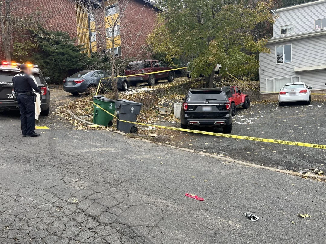 Residential parking area with yellow tape, vehicles, garbage bins and a uniformed person beside a police vehicle.