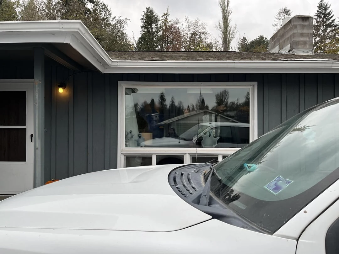 White vehicle hood and windshield in front of a dark-sided house with a large window.