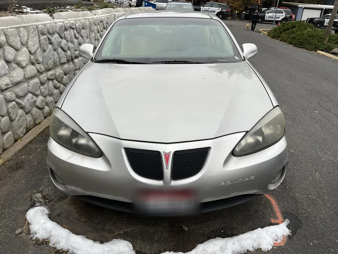 Front of a silver Pontiac sedan beside a stone retaining wall, with a blurred plate and snow near the tires.