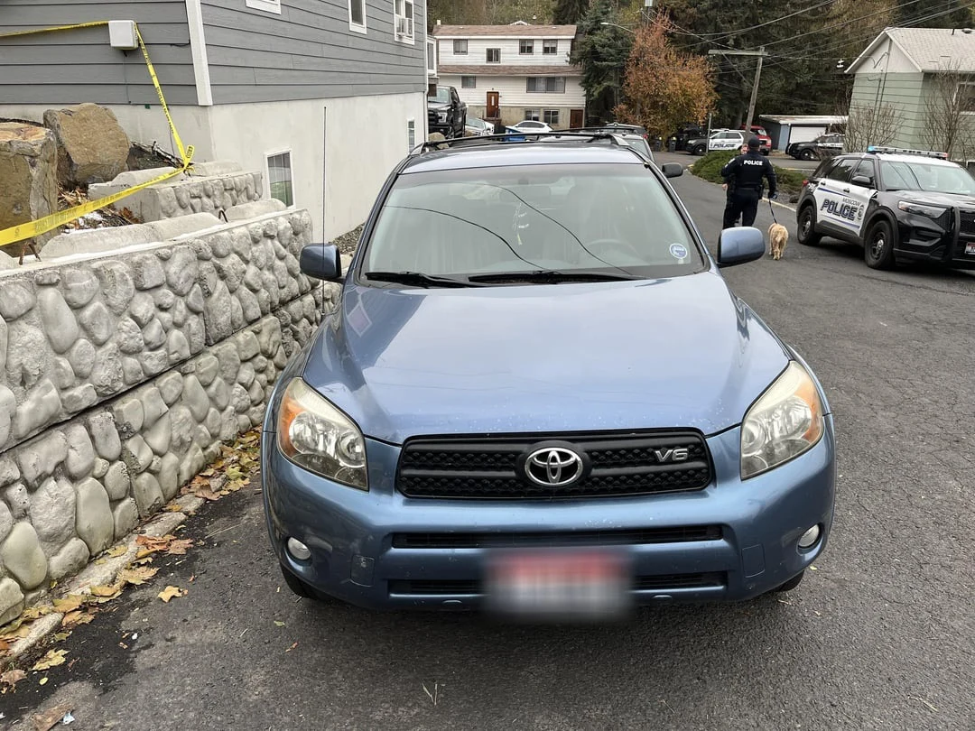 Front of a blue Toyota SUV beside a stone retaining wall, with yellow tape and a police vehicle behind it.