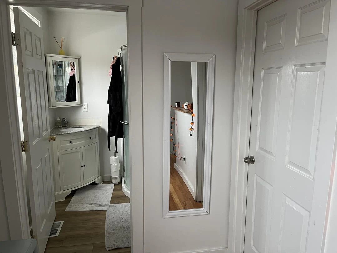 Hallway with a tall wall mirror and white doors beside a bathroom vanity, sink and small rug.