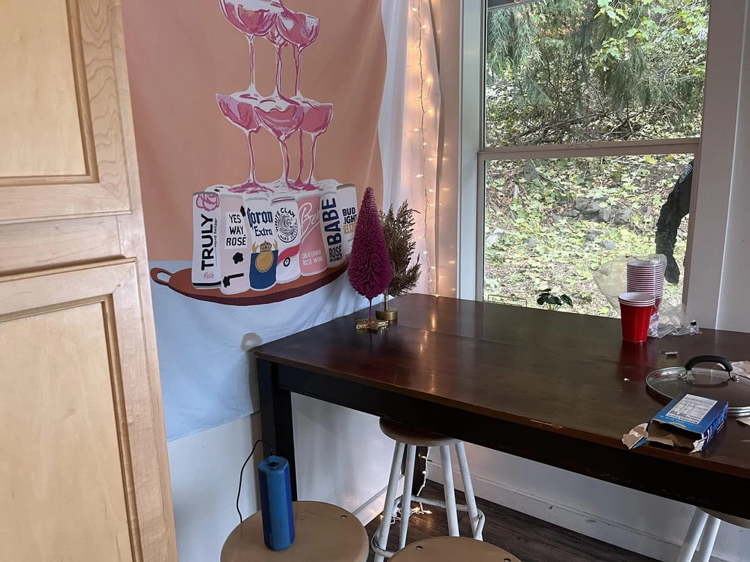 Wooden counter and stools beside a window, red cups and small decorative trees beneath a beverage-themed wall hanging.