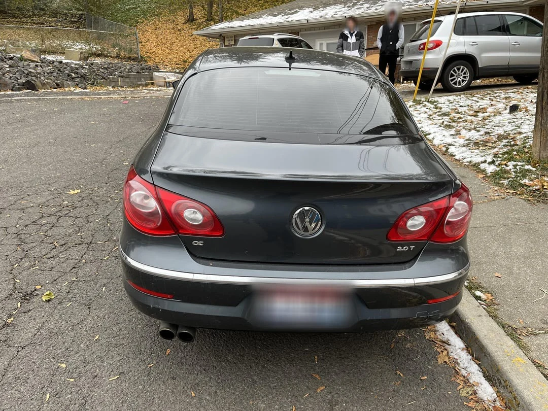 Rear of a black Volkswagen sedan on a residential street, with a blurred plate and blurred people in the background.