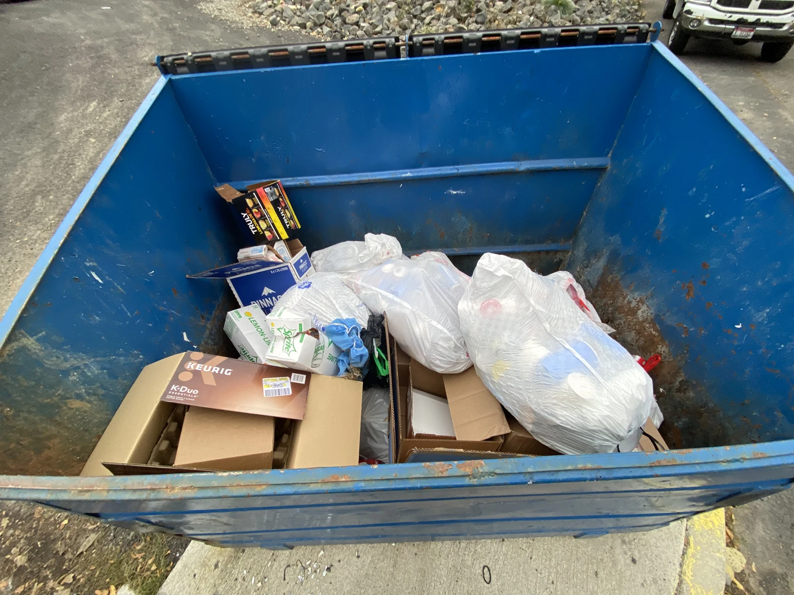 Open blue dumpster containing white trash bags and cardboard boxes.