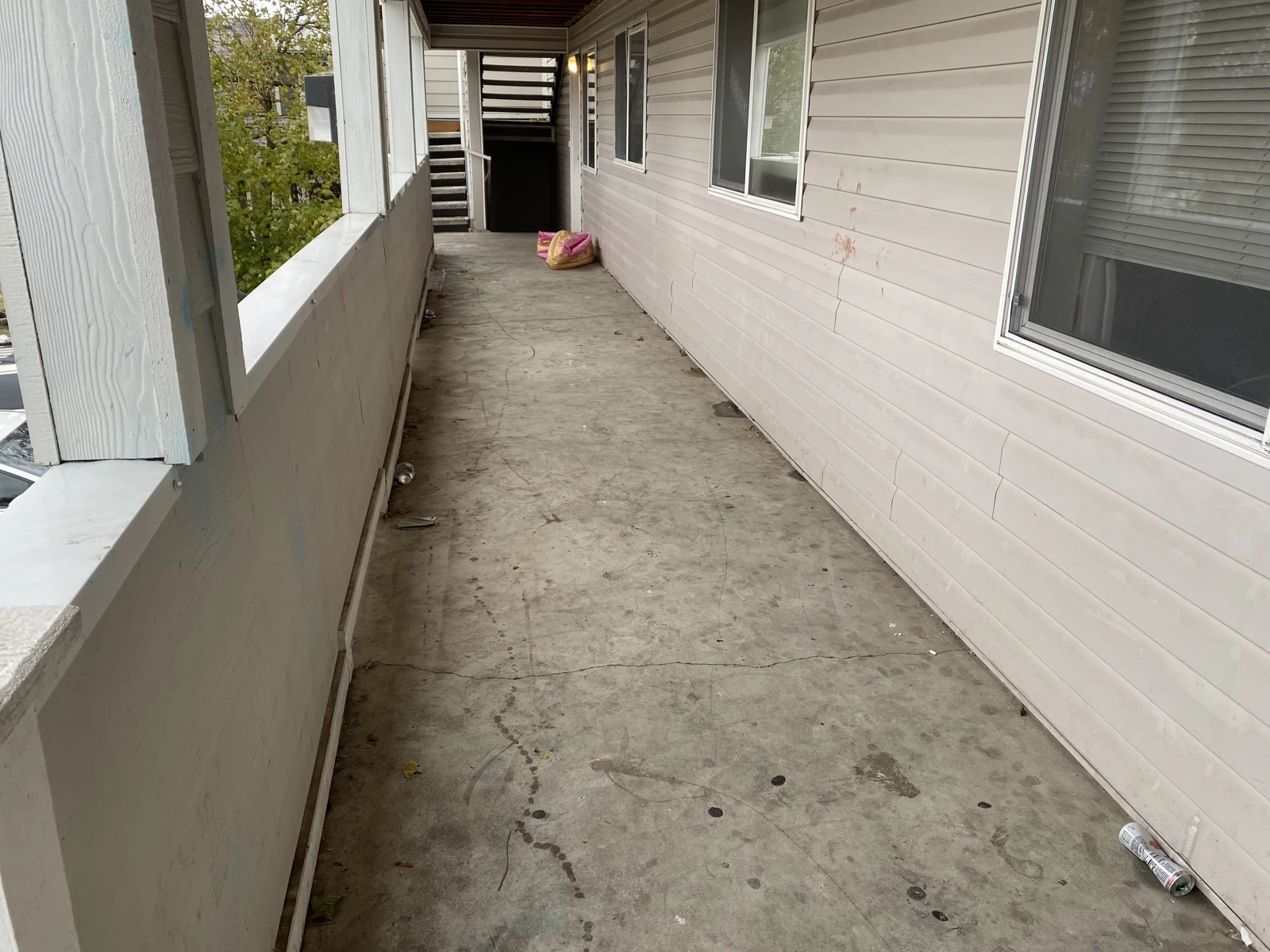 Long exterior apartment walkway with windows, pale siding, concrete flooring and stairs at the far end.