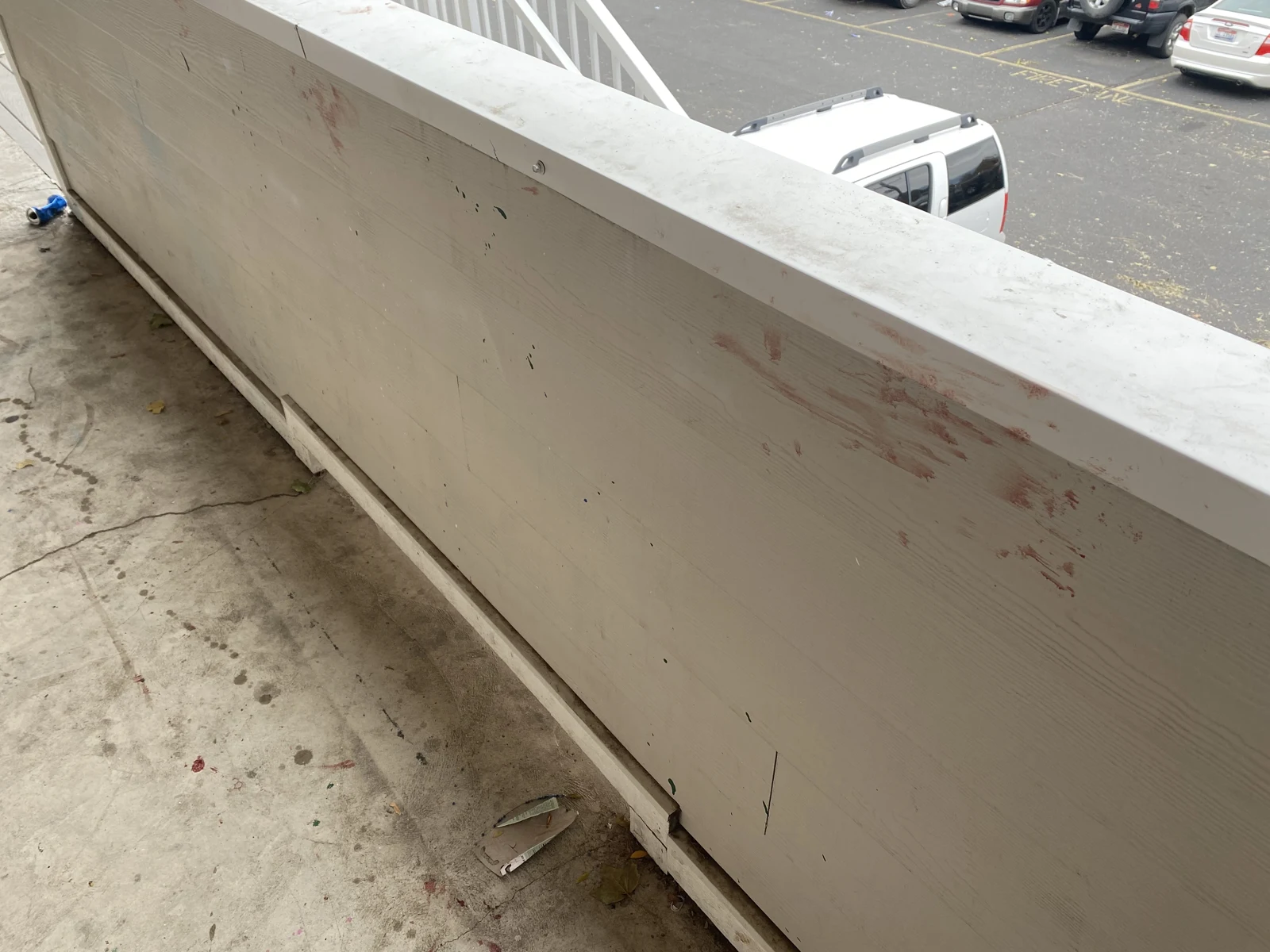 Pale balcony railing with reddish smears, a concrete walkway below and parked vehicles beyond.