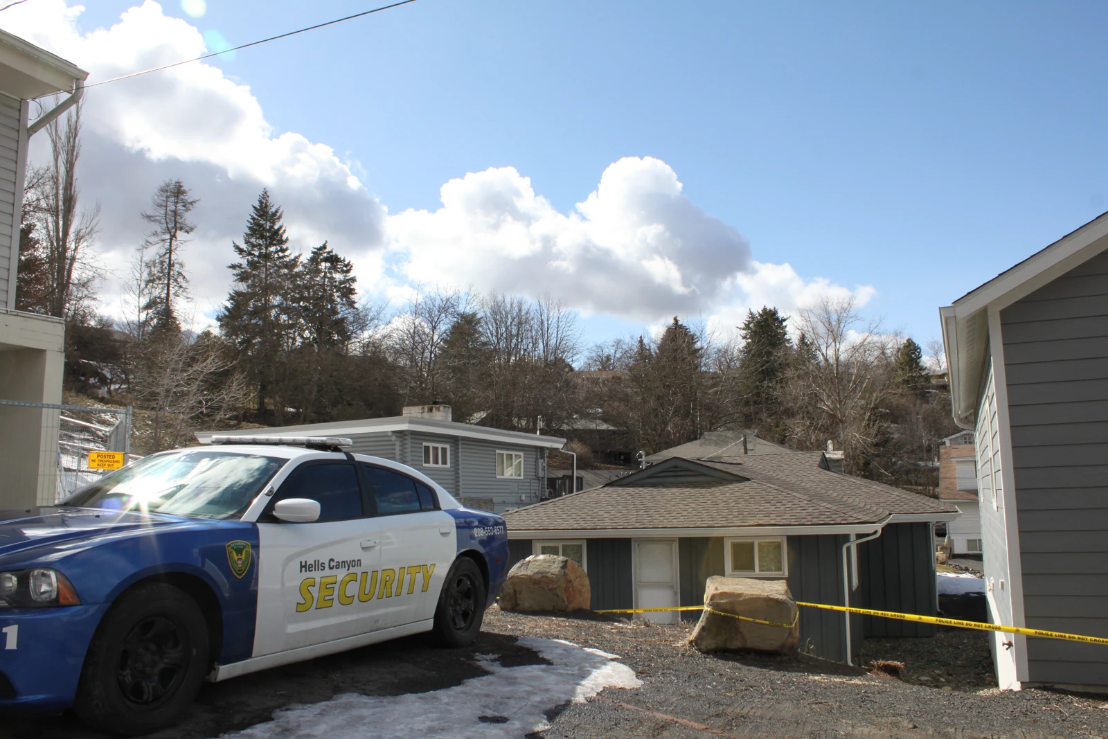 Marked security car beside residential houses, yellow tape and patches of snow under a blue sky.
