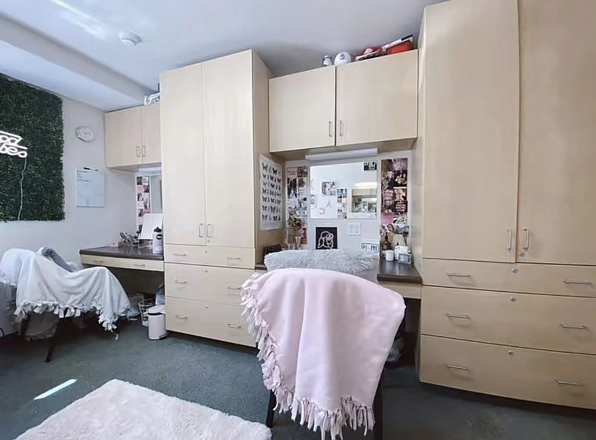 Bedroom study area with light-wood wardrobes, drawers and desks, decorated mirrors, chairs draped with blankets and a light-colored rug.