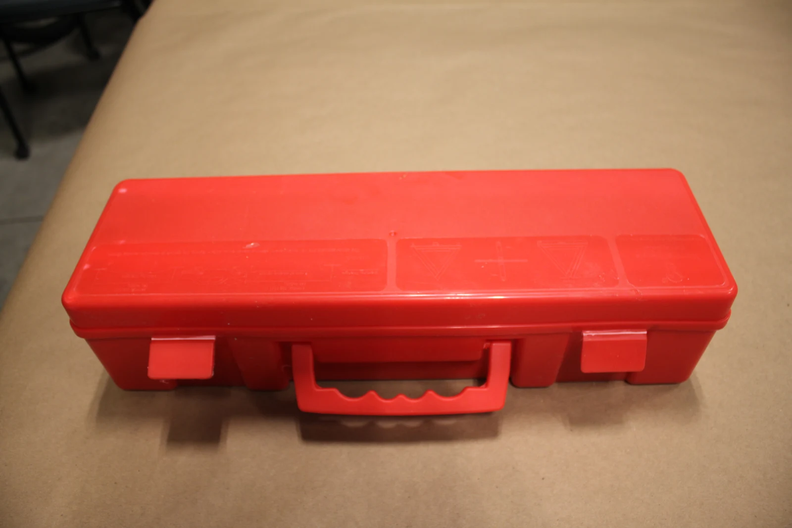 Closed red plastic carrying case with two latches and a handle, resting on brown paper.