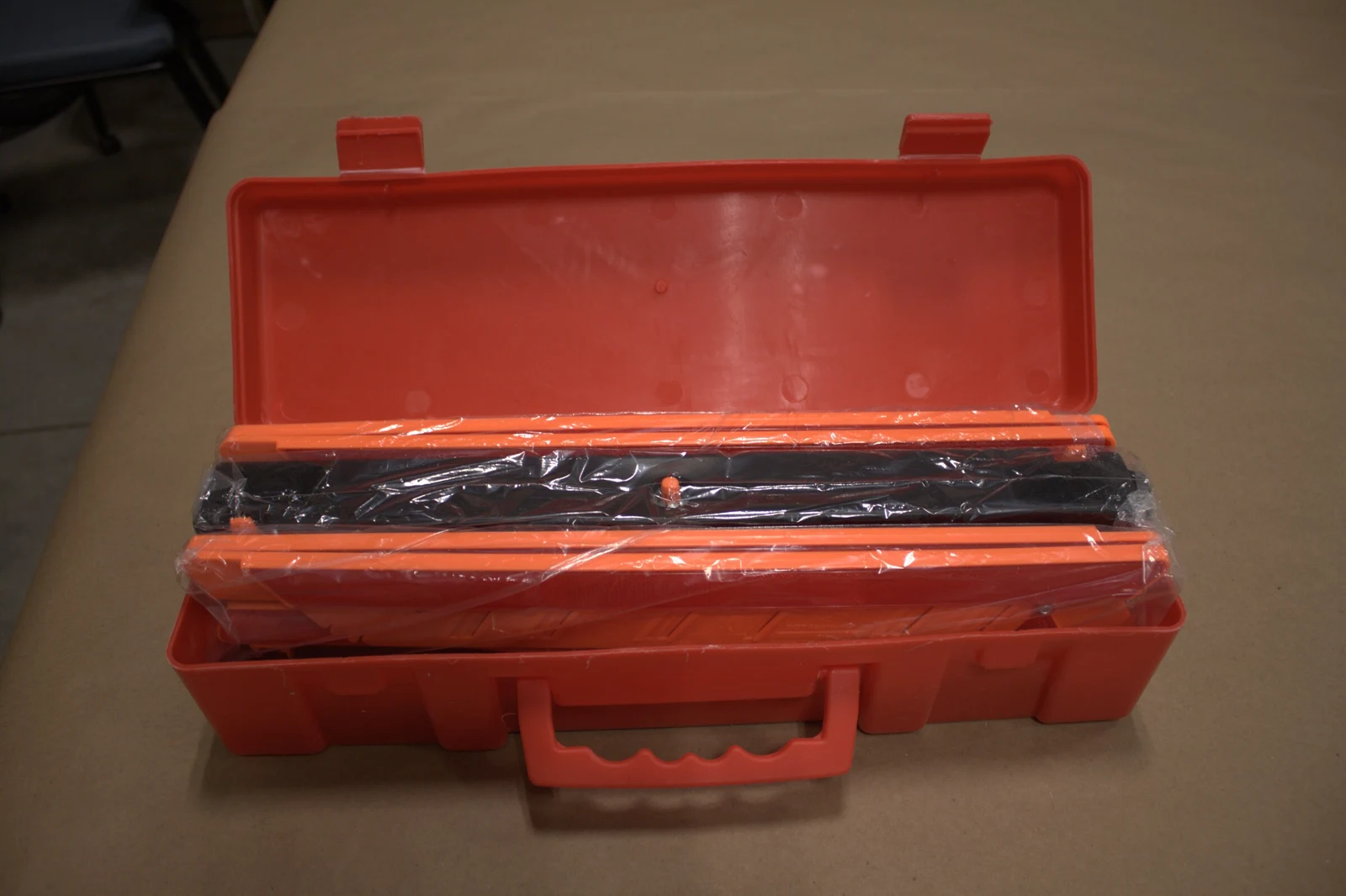 Open red plastic carrying case containing long black and orange objects wrapped in clear plastic.