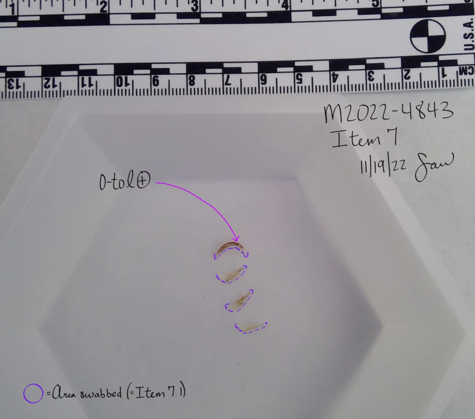 White shallow tray below a measurement scale, with small curved material outlined in purple and handwritten item and swab-area annotations.
