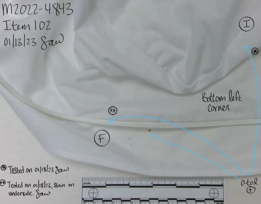 Close photograph of folded white fabric and a seam labeled bottom left corner, with circled marks, blue arrows and a measurement scale.