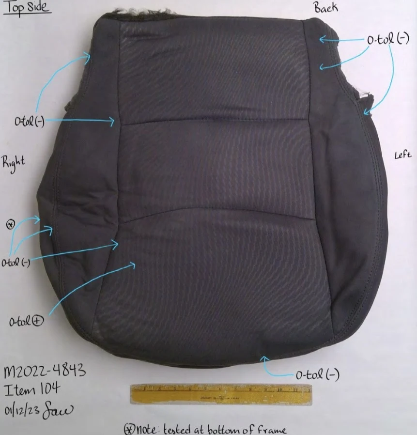 Dark vehicle seat cushion upholstery laid flat beside a ruler, with blue arrows and handwritten top-side testing annotations.