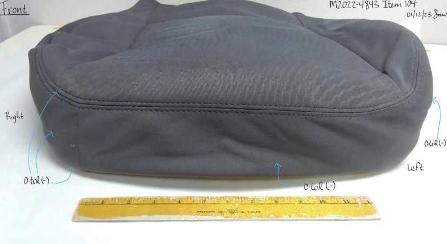 Low front view of dark vehicle seat cushion upholstery beside a yellow ruler, with arrows marking test areas.