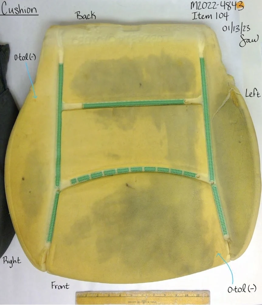 Yellow foam vehicle-seat cushion with green attachment strips and visible discoloration, beside a yellow ruler and test annotations.