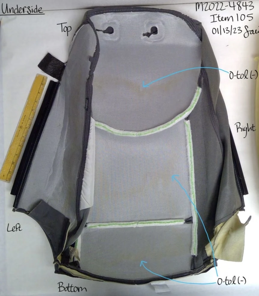 Underside of a removed seat-back cover showing gray backing, curved attachment strips and dark outer edges beside a ruler.