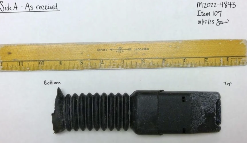 Black vehicle seat-belt buckle assembly with accordion-style covering, photographed intact beside a yellow ruler and labeled Side A as received.