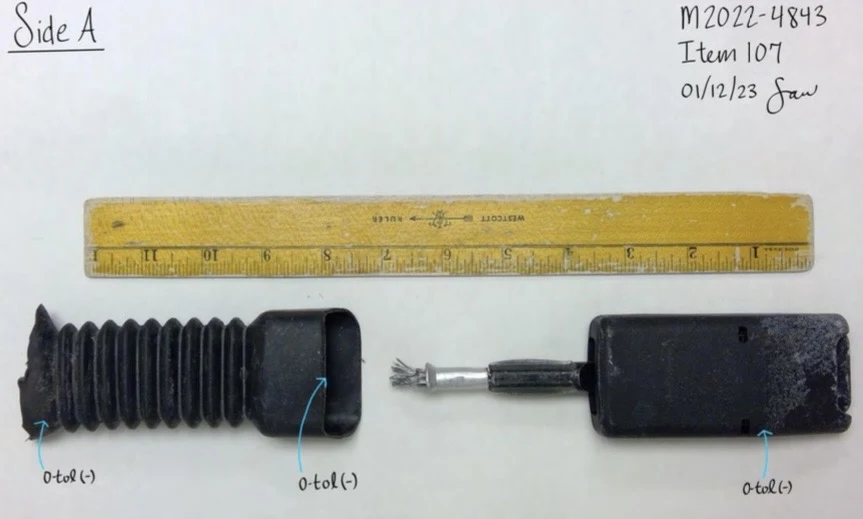 Black seat-belt buckle assembly with accordion covering separated to expose an internal metal cable, photographed beside a yellow ruler.