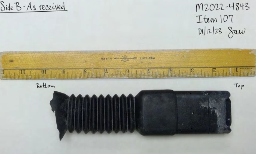 Reverse view of an intact black seat-belt buckle assembly with accordion-style covering, beside a ruler and labeled Side B as received.