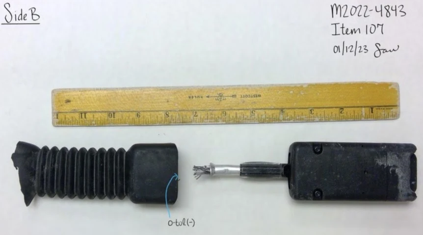 Reverse view of a black seat-belt buckle assembly with covering separated and internal cable visible, beside a ruler.