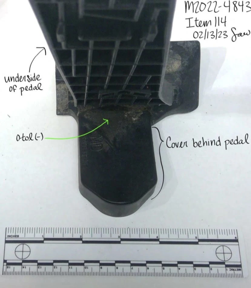 Underside of a raised vehicle pedal showing reinforcing ribs and a cover behind it, with a measurement scale and annotations.