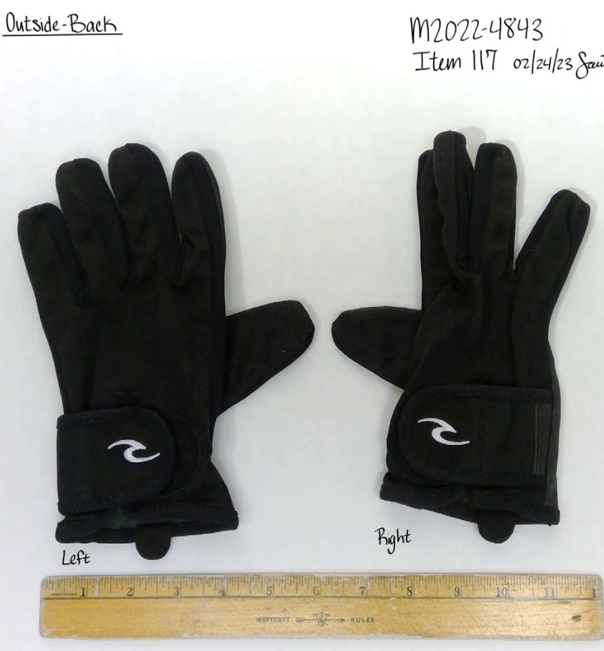 Backs of a pair of black gloves with white curved logos on hook-and-loop wrist straps, beside a yellow ruler.
