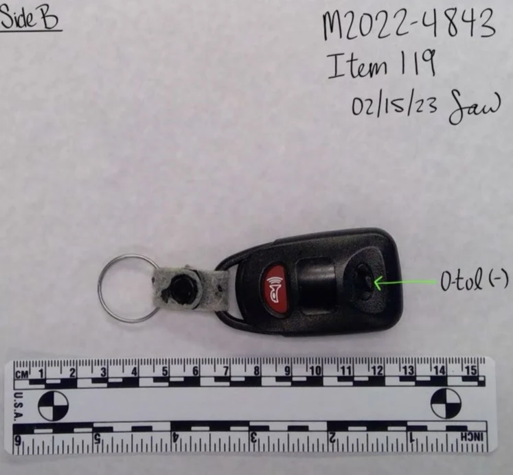 Reverse side of a black vehicle key fob with a red alarm button and metal ring, above a measurement scale.