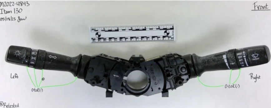 Front of a removed vehicle steering-column switch assembly with two control stalks, symbols and a central circular opening beside a measurement scale.