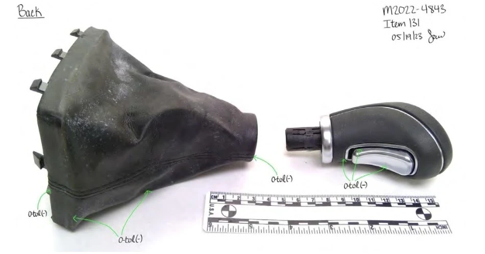 Back view of a gear-shift knob with a silver button and detached black boot, beside a measurement scale.