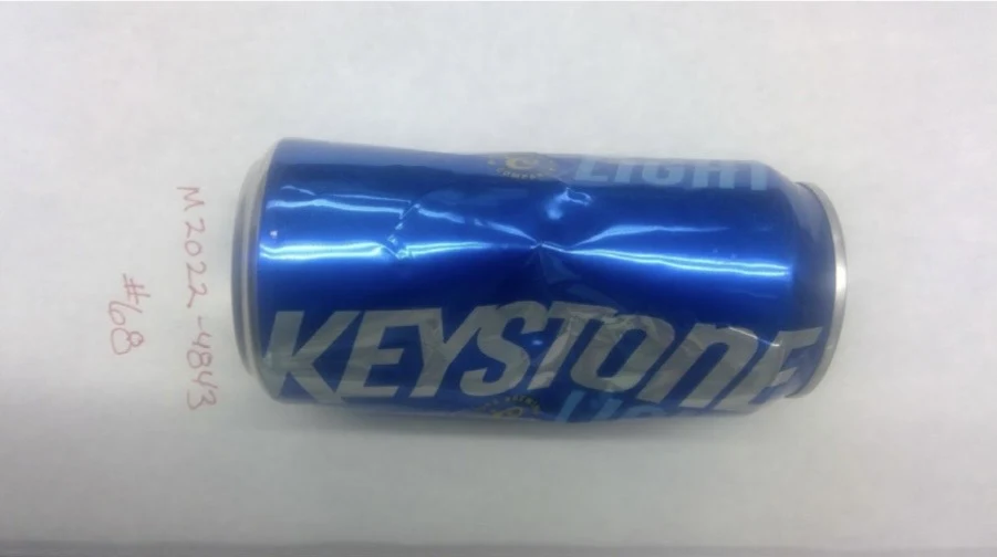 Side of a blue Keystone Light can with a long dent, photographed horizontally above a ruler, labeled item 68.