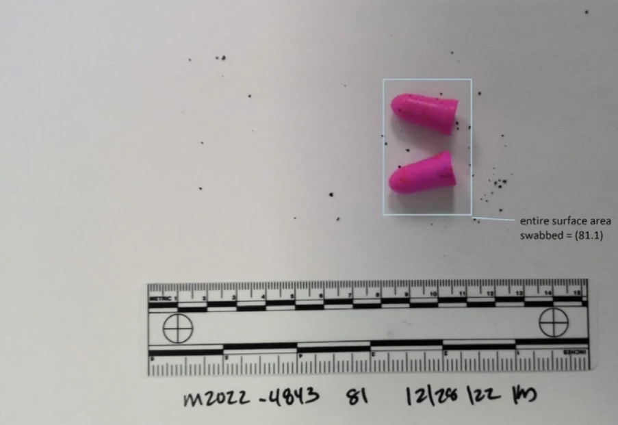 Two bright pink foam earplugs with scattered dark particles, above a measurement scale and inside a swab-area outline.