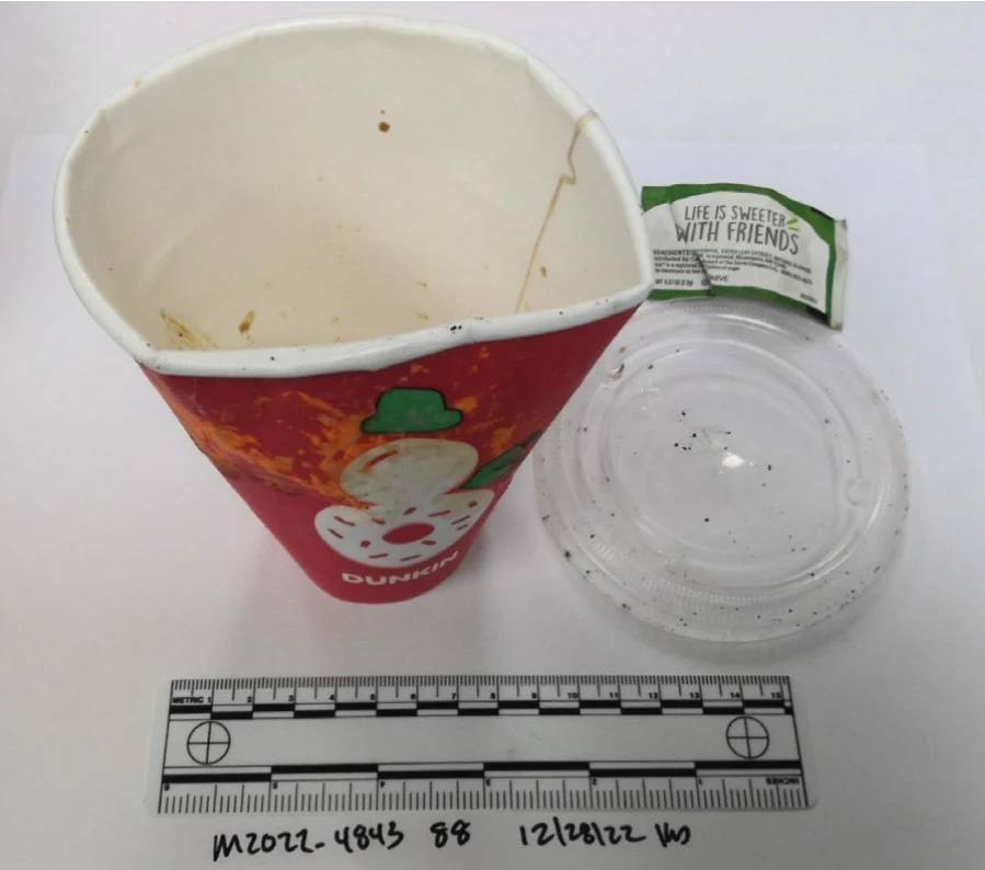 Used red Dunkin paper cup with a snowman design, clear plastic lid and small green wrapper, above a measurement scale.