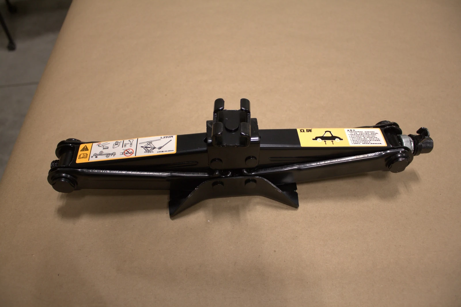 Folded black scissor jack lying horizontally on brown paper, with warning labels visible.