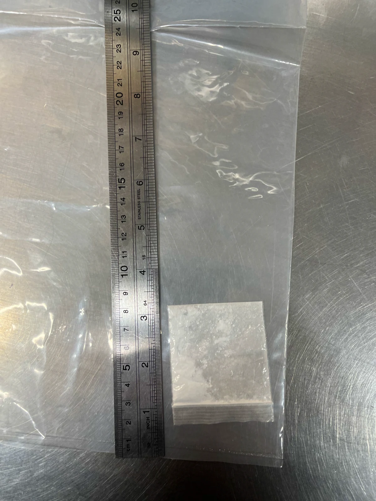 Clear plastic bag containing a smaller bag with pale granular material, beside a metal ruler on a metal surface.