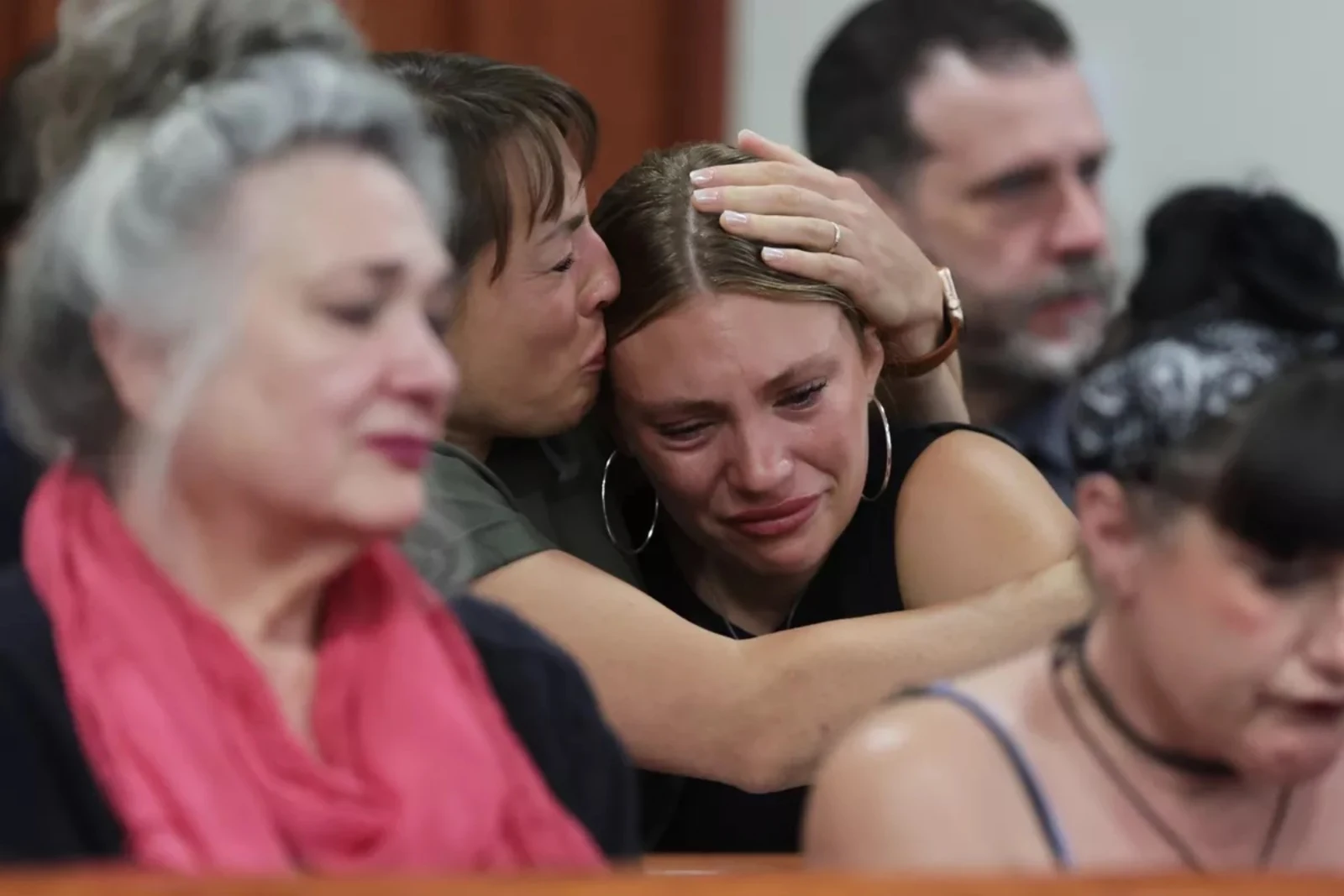 Seated people in a courtroom; one person embraces and kisses the head of another person who appears tearful.