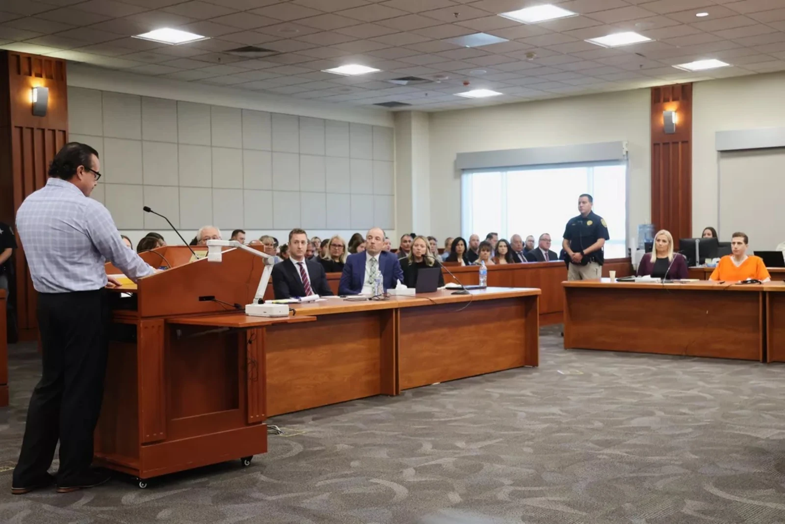 Wide courtroom view with a person at a wooden lectern, seated attendees, counsel tables, microphones and a person in orange clothing.