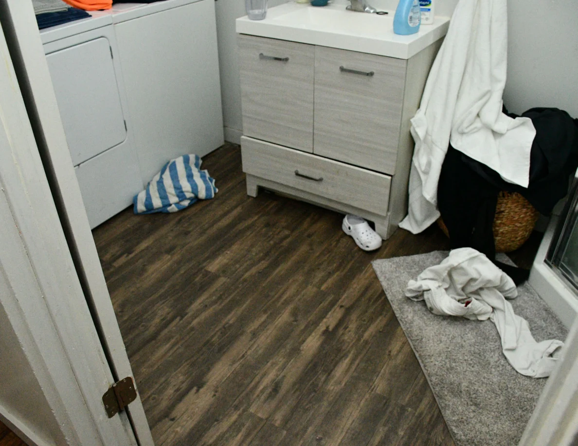 Bathroom vanity, laundry appliances, towels and footwear on wood-pattern floor.