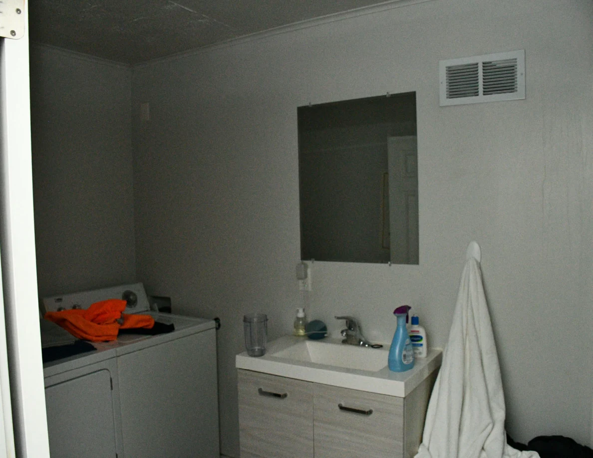 Bathroom sink and mirror beside laundry appliances, towels and a glass shower enclosure.