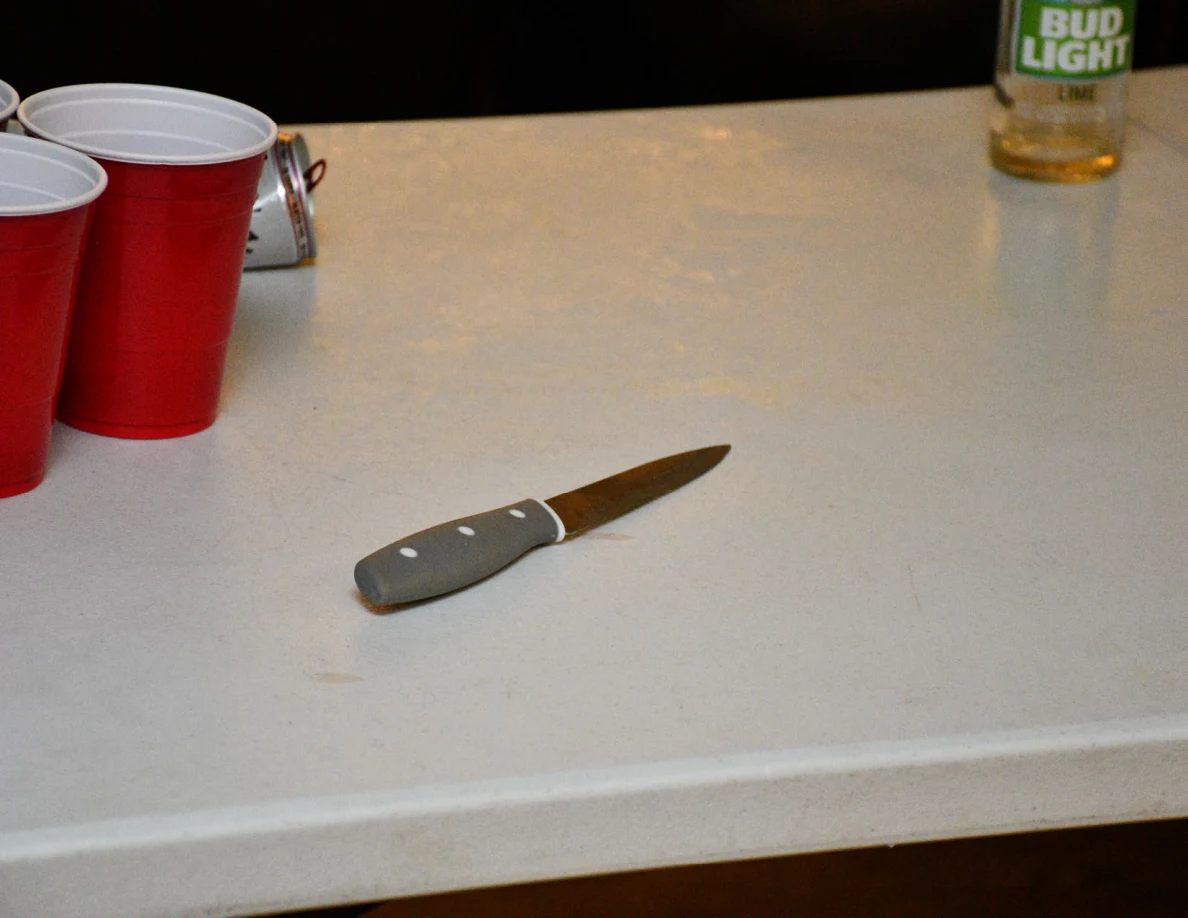 Close view of a small knife on a white folding table beside red cups and a bottle.