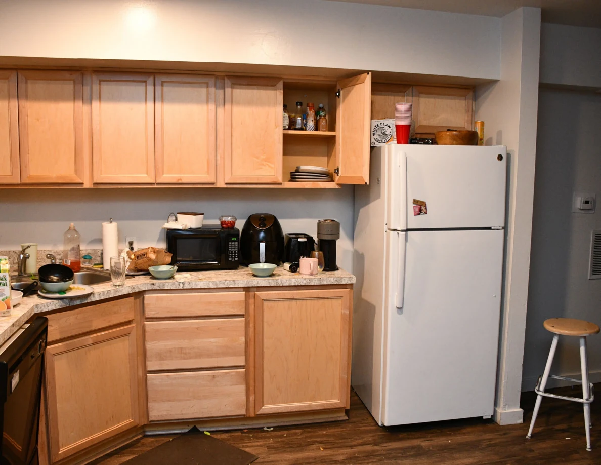 Kitchen cabinets and countertop with microwave, kettle, dishes and refrigerator.