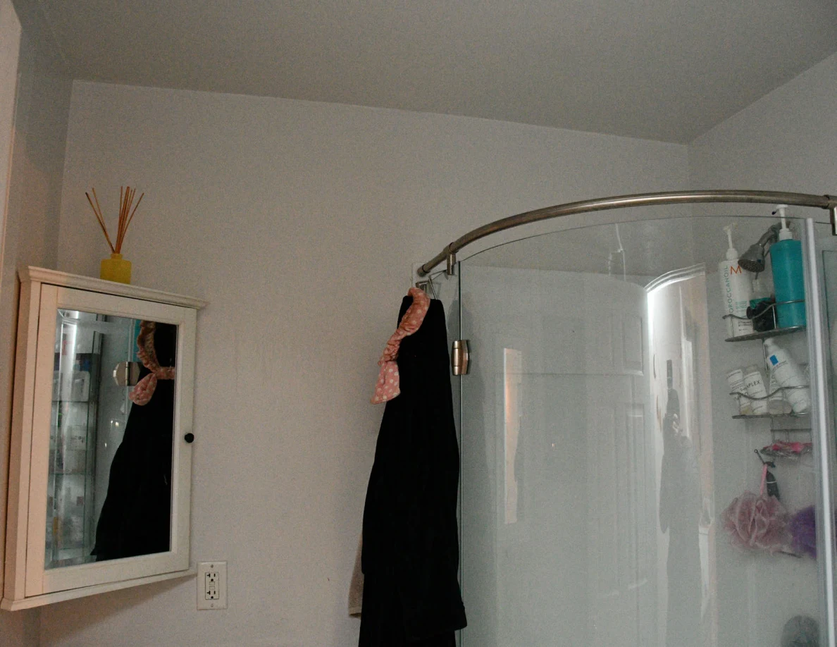 Bathroom mirror and curved shower enclosure with hanging dark clothing.
