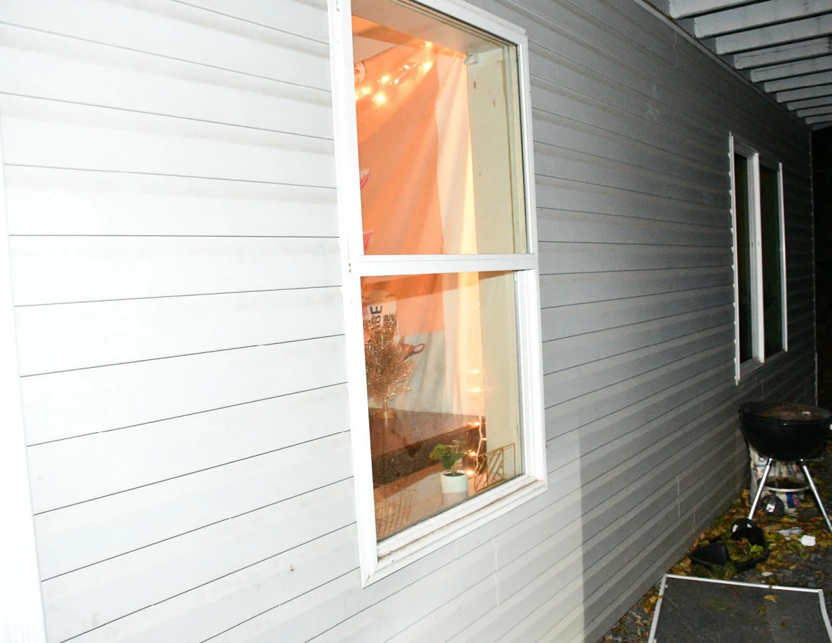Lit window in exterior siding with kettle barbecue grill nearby.