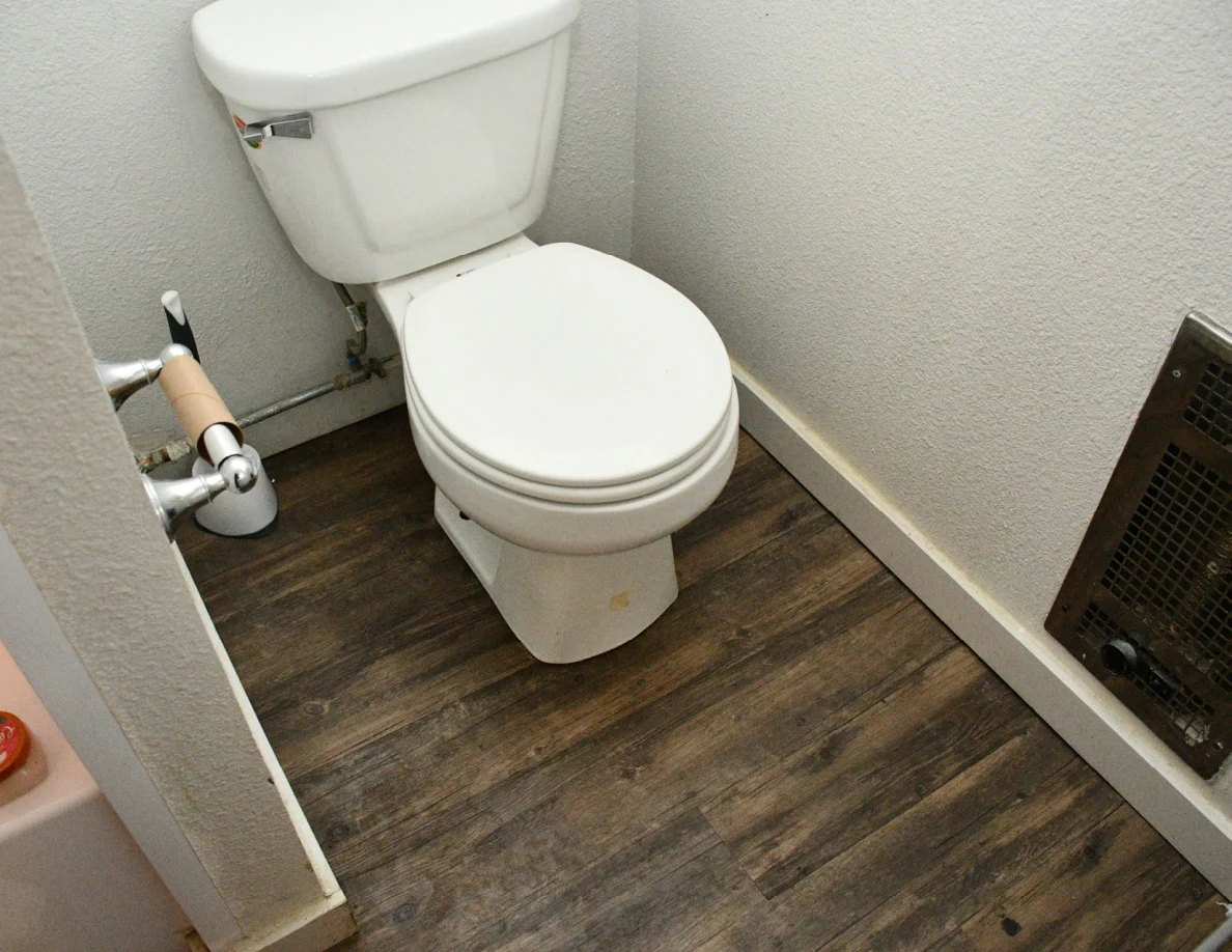 Closed toilet beside wall heater on wood-pattern bathroom floor.
