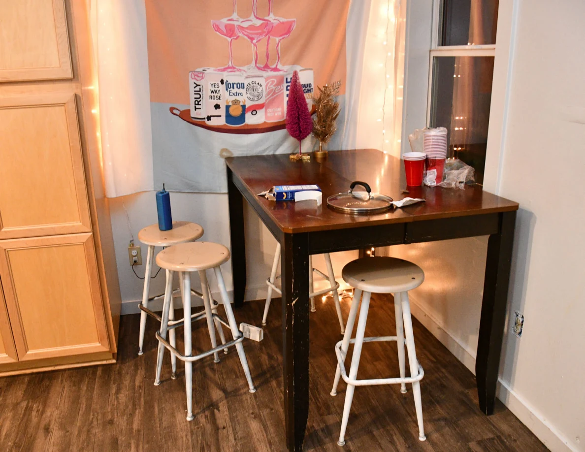 Tall wooden table and stools with cups and a pan lid beside kitchen cabinets.