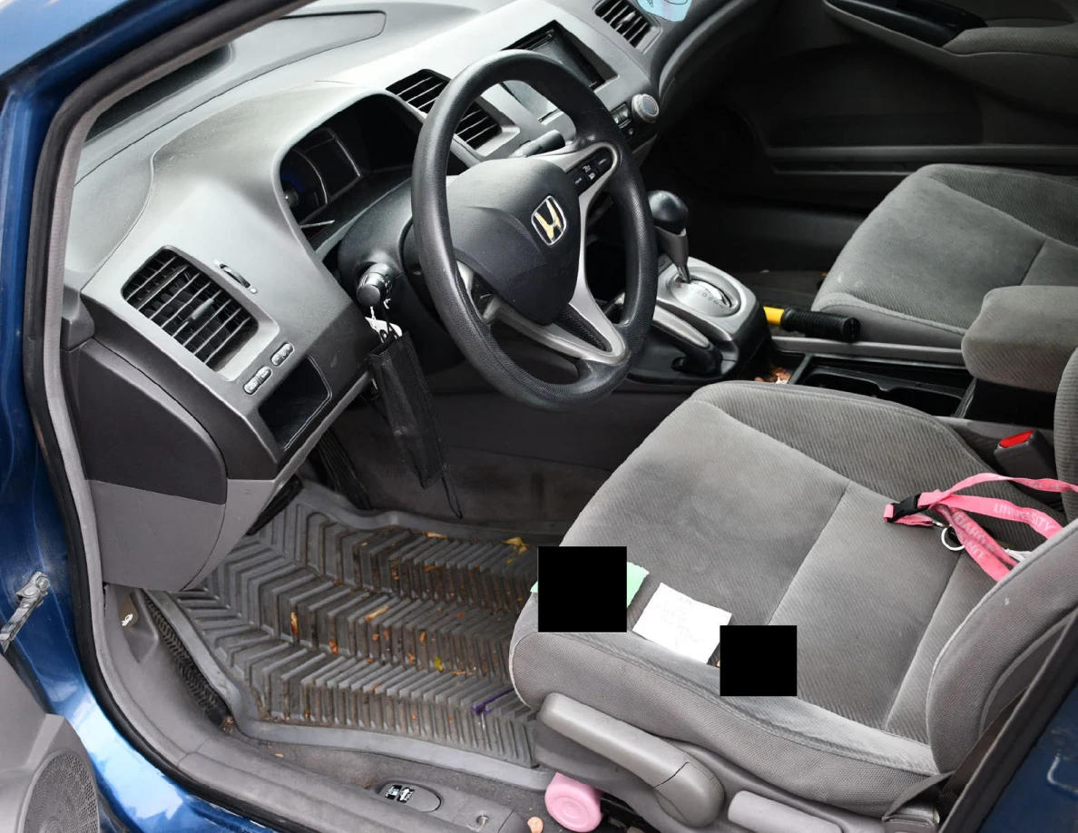 Car interior with steering wheel, dashboard and fabric front seats; items partly redacted.