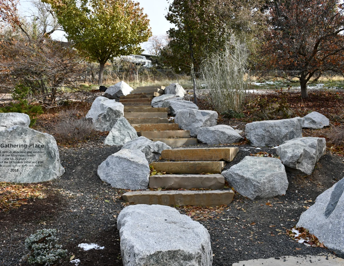 Outdoor stone staircase bordered by large gray rocks and shrubs, with an engraved Gathering Place stone at the side.