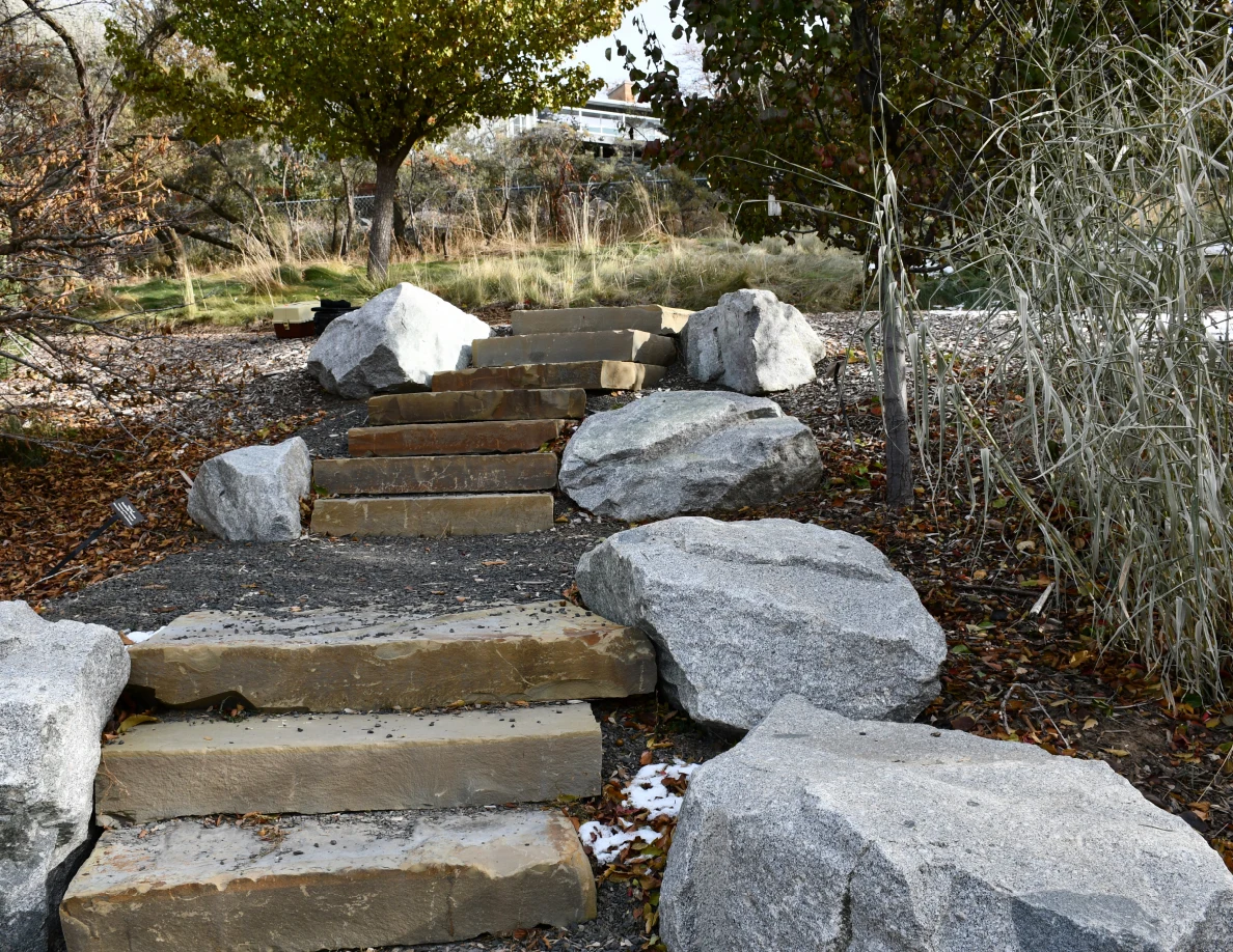 Stone steps and gravel landings among large gray rocks, fallen leaves and tall grasses.