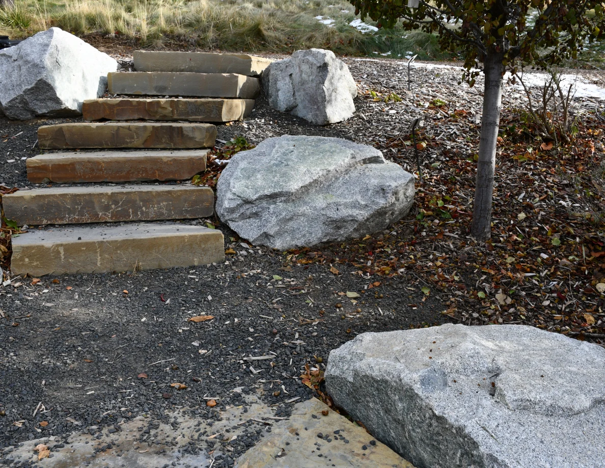 Closer view of stone steps beside gray boulders and a tree, with fallen leaves on gravel.