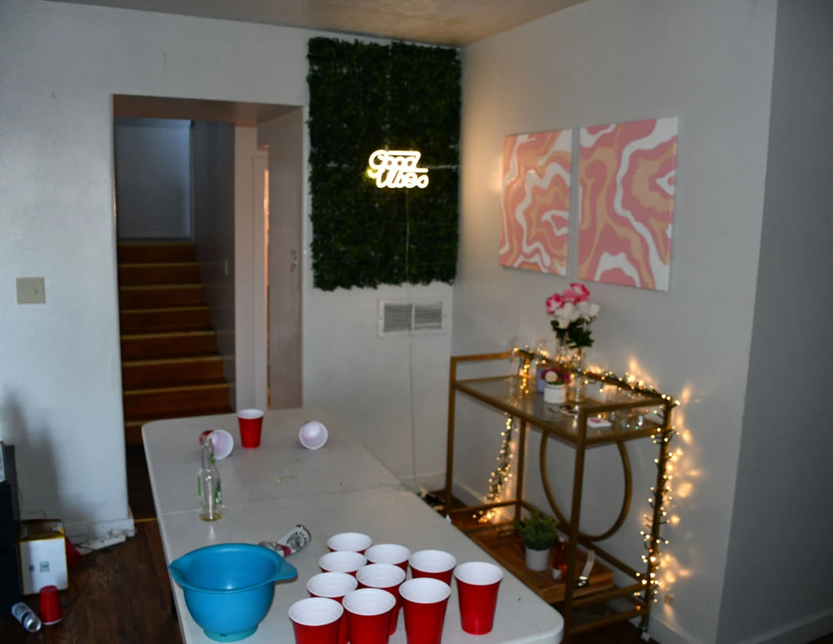 White cup-game table with red cups, a blue bucket and bottle beside a string-lit glass console, abstract art and an illuminated Good Vibes sign.